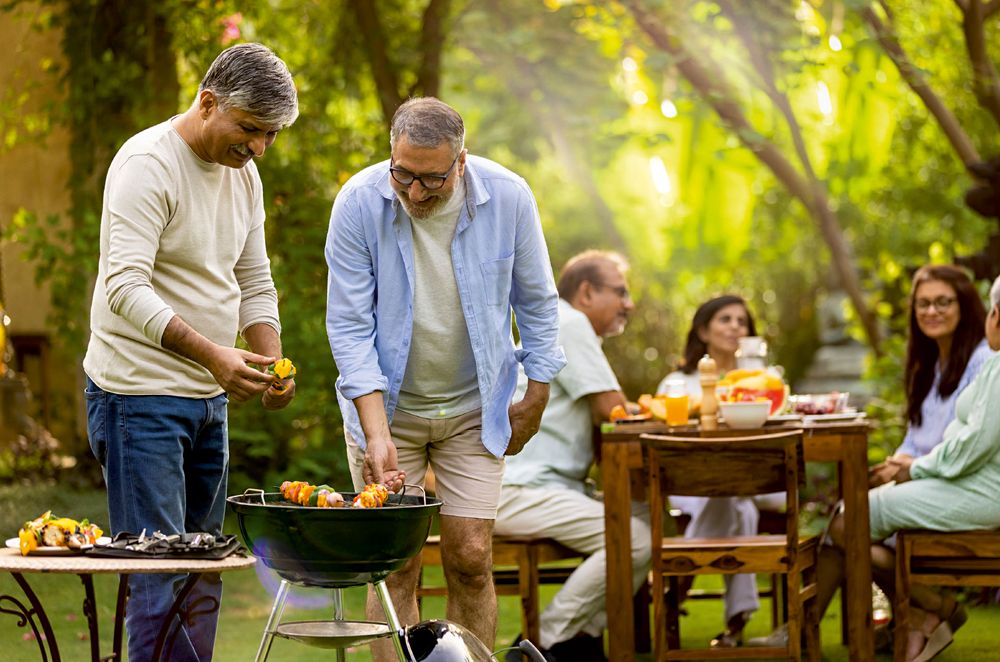 A family gathered around a table on a lawn while two men barbecue